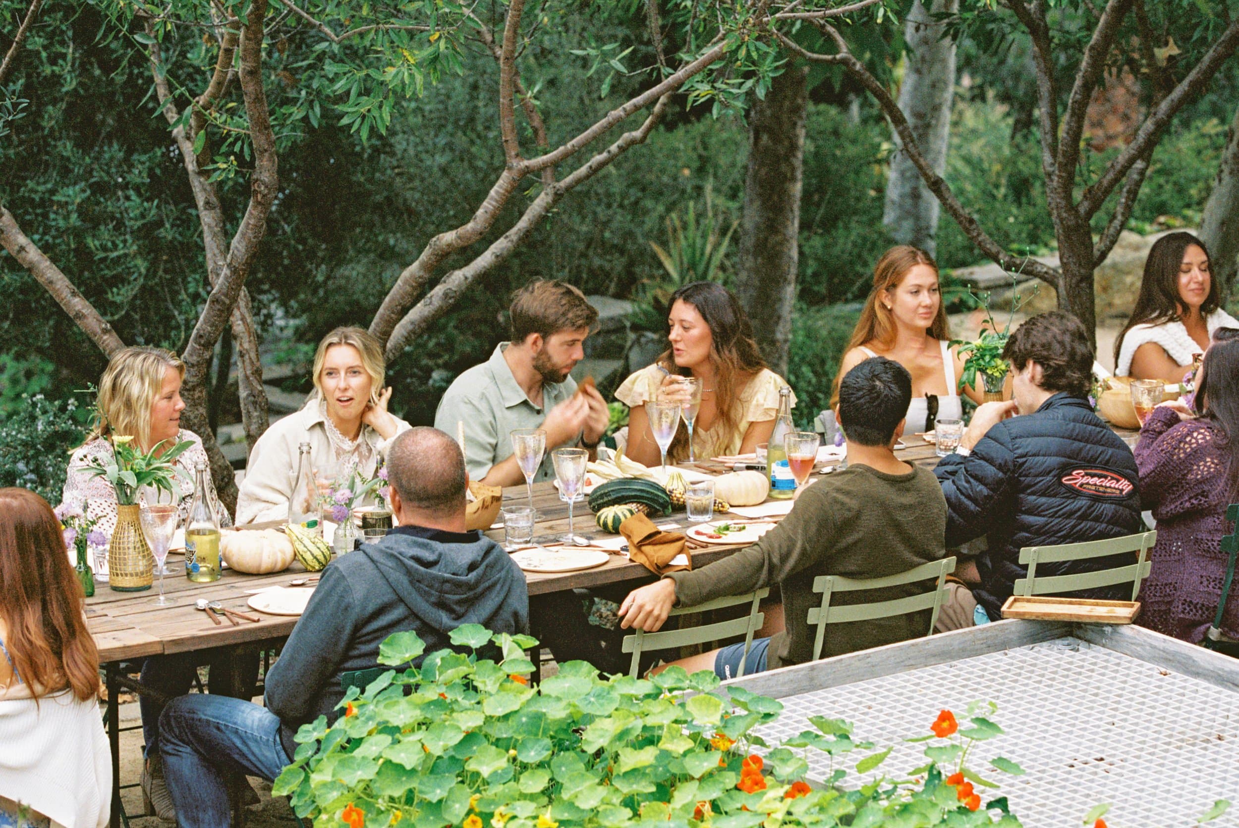 A long outdoor table under canopy trees, set with linen, garden flowers, and seasonal squash — guests in conversation over a Lighthearted Kitchen meal.